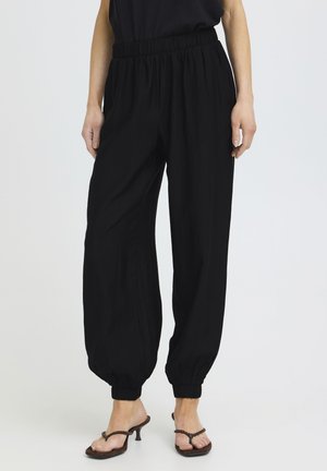Mujer con pantalones jogger de cintura elástica y puños elásticos en color negro, combinados con sandalias marrones de tacón, de pie frente a un fondo blanco.