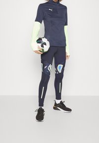 Haut de sport à manches longues bleu marine avec des accents vert fluo, associé à un legging ajusté avec des motifs abstraits et des bandes réfléchissantes. Tient un ballon de soccer.