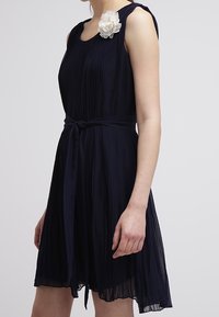 Laivastonsininen laskostettu mekko, jossa on tiukka vyötärö, pyöreä kaula-aukko ja kangaskukkakoriste vasemmassa olkapäässä. Pehmeä ja kevyt kangas.