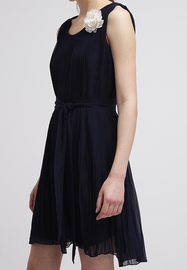 Laivastonsininen laskostettu mekko, jossa on tiukka vyötärö, pyöreä kaula-aukko ja kangaskukkakoriste vasemmassa olkapäässä. Pehmeä ja kevyt kangas.