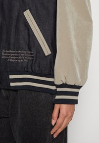 Person som bär en svart och beige varsityjacka med randiga ribbade muddar och midja, stående med en hand avslappnad vid sin sida.