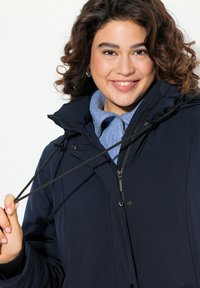 Navy blue insulated jacket with a zippered front, adjustable drawstrings, and large hood. Paired with a light blue collared shirt beneath.
