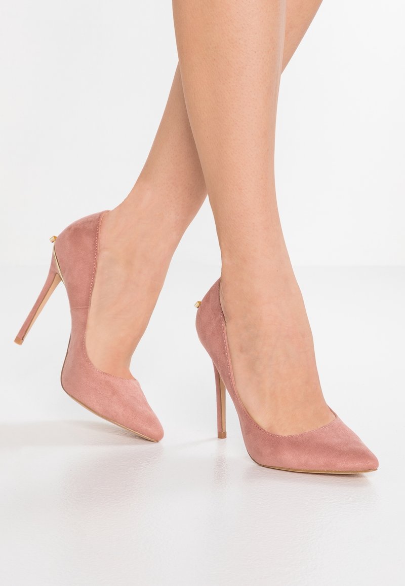 Jambes portant des escarpins en daim rose poudré à bout pointu avec talons aiguilles sur un sol blanc.