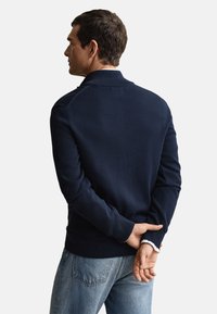 Marineblå strikkegenser med krage, har ribbestrikkede mansjetter og linning, brukt over lyseblå jeans, sett bakfra.