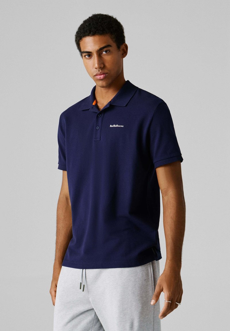 Junger Mann trägt ein marineblaues Polohemd mit kleinem "Red Bull Racing"-Logo, kombiniert mit hellgrauen Jogginghosen vor neutralem Hintergrund.