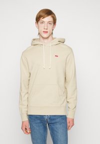 Beige hettegenser med frontlommedesign, justerbar hette med snøring og liten rød logo på brystet. Mykt stoff og avslappet passform.