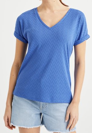 Blaues V-Ausschnitt-T-Shirt mit kurzen aufgerollten Ärmeln und einer strukturierten Fischgrätmuster-Optik. Kombiniert mit hellen Denim-Shorts.