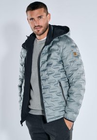 Puffer jacket in light gray with a black hood, zippered front, and textured pattern. Features a logo patch on the left sleeve.