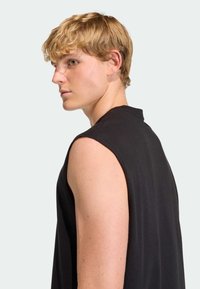 Čierny top bez rukávov z mäkkej tkaniny, s vysokým výstrihom a voľným strihom. Modelka je zobrazená z profilu, zvýrazňujúc detail svalnatého ramena.