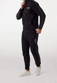 Schwarzer Trainingsanzug mit Reißverschlussjacke, Fronttasche und passenden Hosen. Mit weißem Logo und seitlichen Streifen. getragen mit weißen Sneakers.