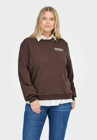 Žena oblečená v hnědém svetru s nápisem "ESPRESSO YOURSELF", pod ním má bílou košili a modré džíny, stojící před jednobarevným pozadím.