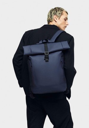 Sac à dos roll-top bleu marine fabriqué dans un matériau durable avec fermeture à boucle noire, poches latérales et marquage en relief sur le panneau avant.