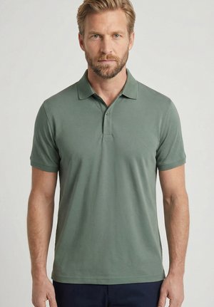 Blonde man met baard, gekleed in een groen polo met korte mouwen, staand tegen een effen lichtgrijze achtergrond.