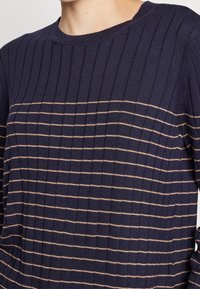 Granatowy sweter prążkowany w poziome beżowe paski, z okrągłym dekoltem i teksturowanym splotem. Idealny do noszenia na warstwy w chłodniejsze dni.