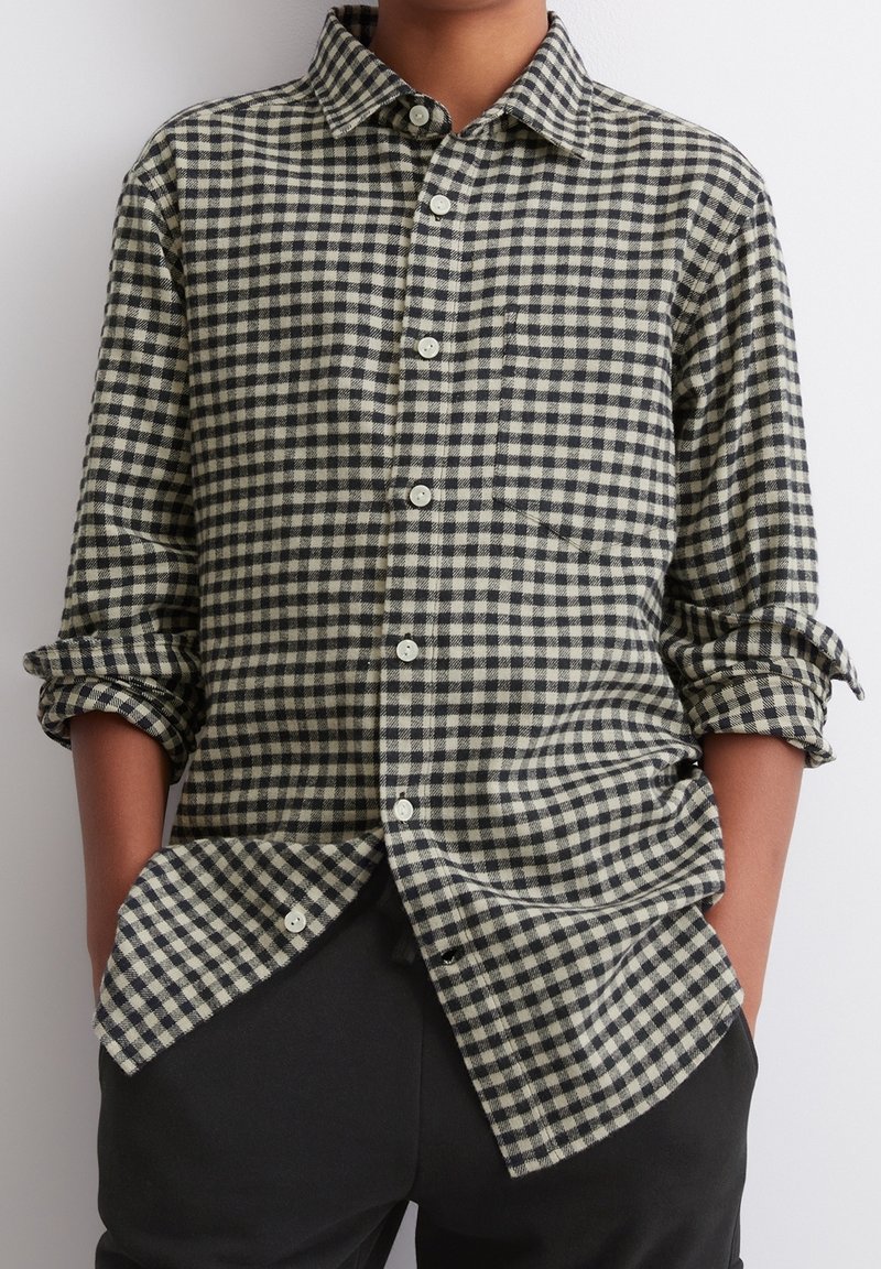 Personne portant une chemise à carreaux noir et blanc boutonnée avec les manches retroussées et un pantalon noir, mains dans les poches sur fond uni.
