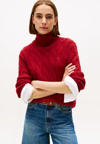 Suéter de punto rojo con cuello alto, diseño de trenzas texturizadas y puños de camisa blanca. Combinado con jeans de mezclilla azul y un cinturón marrón.