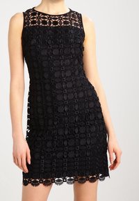 Svart ärmlös klänning med intrikata cirkulära spetsmönster, en fast halsringning och vågig nederkant. Tillverkad av texturerat tyg.