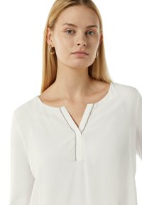 Weiße Bluse mit langen Ärmeln und V-Ausschnitt, der mit einem silbernen Akzent verziert ist. Weicher, glatter Stoff mit lässiger Passform.