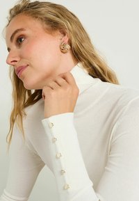 Suéter blanco de cuello alto con textura y botones redondeados en los puños. Pendientes de aro dorados visibles en un perfil inclinado.