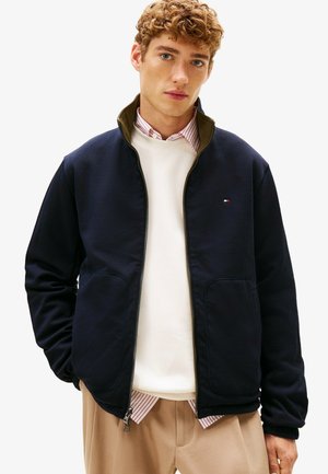 Jeune homme aux cheveux bouclés, portant une veste zippée bleu marine sur un sweat-shirt blanc et un pantalon beige, debout sur un fond blanc.