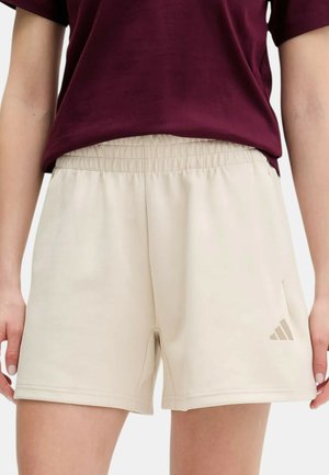 Persona che indossa pantaloncini beige con vita elastica con logo Adidas sulla gamba sinistra e una maglietta a maniche corte color bordeaux, in piedi con le braccia rilassate.