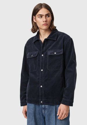 Jonge man met schouderlang haar die een donkerblauw corduroy jasje met knopen en losse blauwe spijkerbroek draagt, kijkt opzij.