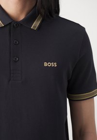 Polo bleu marine avec trois boutons, col rayé doré, et logo "BOSS" doré sur la poitrine, tissu texturé, manches courtes.