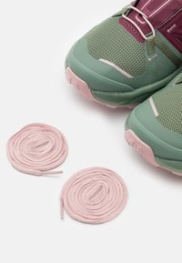 Baskets de sport vertes avec une tige en tissu mesh et des semelles en caoutchouc, accompagnées de deux lacets ronds roses. La vue en gros plan met en valeur la texture et le contraste de couleurs.