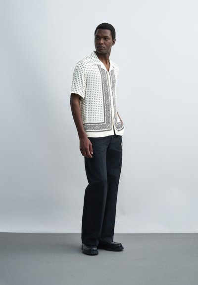 Άνδρας που φοράει λευκό κοντομάνικο πουκάμισο με σχέδια, μαύρο παντελόνι με φαρδιά πόδια και μαύρα παπούτσια, στέκεται σε απλό ανοιχτό γκρίζο φόντο.