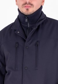 Giacca blu navy con colletto alto, chiusura con zip e bottone, e due tasche frontali con zip. Tessuto texturizzato con una finitura liscia.