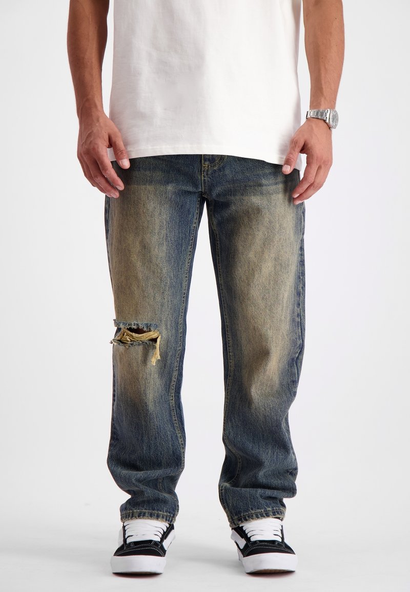 Blauwe denim jeans met een losse pasvorm, met een gescheurde kniedetail, lichte vervaging en een recht toelopende pijp, gecombineerd met zwart-witte sneakers.