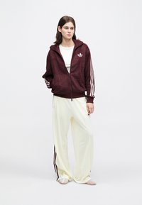 Mulher vestindo uma jaqueta burgundy com fecho éclair e riscas brancas, calças largas brancas com riscas burgundy e sapatos bege, de pé com uma mão no bolso.