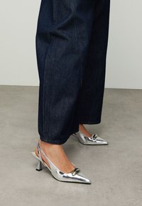 Escarpins à bride arrière argentés avec un bout pointu, une texture brillante, un petit talon carré et un accent circulaire à l'avant. Portés avec un pantalon en jean foncé.