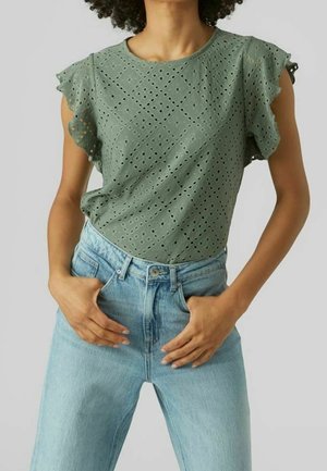Personne portant un chemisier vert à manches courtes ajouré, rentré dans un jean taille haute bleu clair, les mains reposant près des poches.