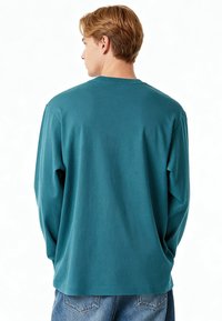 Jeune homme aux cheveux blonds courts, portant une chemise à manches longues bleu sarcelle et un jean bleu, de dos sur un fond blanc uni.