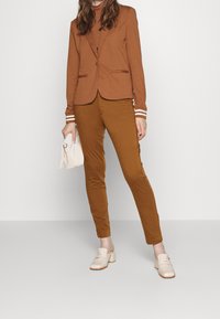 Blazer brown aderente con polsini a righe, abbinato a pantaloni slim-fit marroni e mule con tacco a blocco beige. Modella che tiene una pochette bianca.