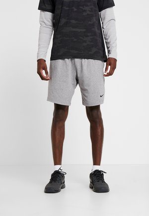 Athlète portant un short gris Nike, des baskets noires, des chaussettes blanches, un t-shirt noir à motif camouflage sur des manches blanches longues, debout sur fond blanc.