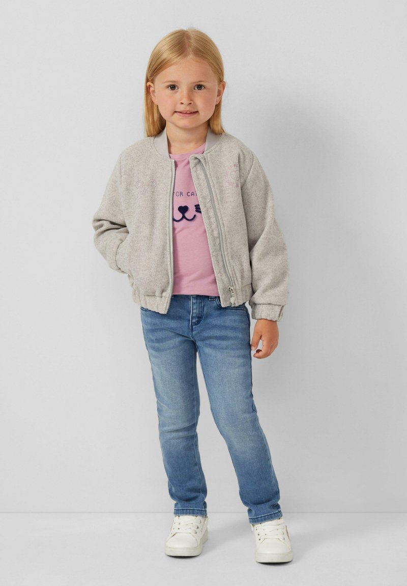 Grau Bomberjacke mit gerippten Bündchen, rosa T-Shirt mit Katzenmotiv, blaue Jeans und weiße Sneakers, stehend vor einem neutralen Hintergrund.