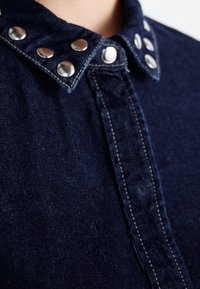 Camicia in denim di un blu scuro, con bottoni a scatto argentati sul colletto e dettagli di cucitura visibili lungo la parte frontale.
