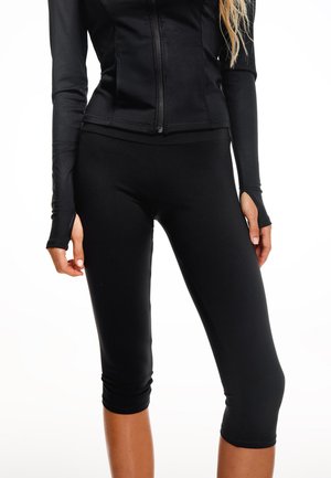 Femeie purtând un top negru cu mâneci lungi și fermoar și colanți negri scurtați, stând cu mâinile relaxate pe lângă corp pe un fundal alb.