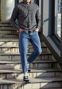 Camicia a scacchi nera e grigia, jeans azzurri chiari e sneakers nere con dettagli bianchi, in piedi su una scaletta con gradini in cemento.