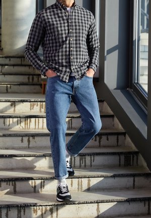Chemise à carreaux noire et grise, jean bleu clair et baskets noires avec accents blancs, debout sur un escalier à marches en béton.