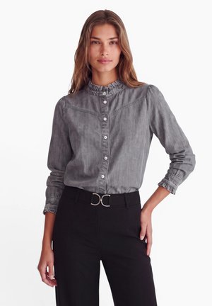 Jeune femme portant un chemisier gris boutonné avec un col à volants et un pantalon noir avec une boucle de ceinture argentée, debout avec une main sur la hanche.