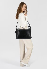 Bolso de mano de piel negra con textura suave, correa de hombro única y forma rectangular. Incluye una etiqueta y es llevado por un modelo.