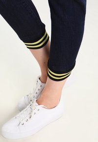 Donkerblauwe denim joggers met zwarte en gele geribbelde boorden, gedragen met witte sneakers met een gestructureerde zool en veters.