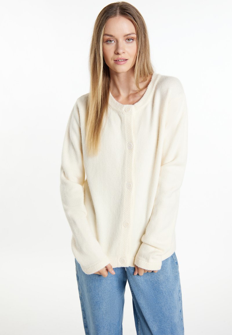 Crème kleurige cardigan, zachte textuur, ronde halslijn, knoopsluiting, lange mouwen, gecombineerd met lichtblauwe denim broek.