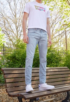 Personne portant un t-shirt blanc Levi's, un jean bleu clair et des baskets blanches, debout sur un banc en bois dans un parc avec des arbres et de la verdure en arrière-plan.