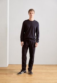 Bărbat stând în interior pe un podea din lemn, purtând o cămașă lungă de culoare închisă, pantaloni închiși și adidași negri cu dungi albe.