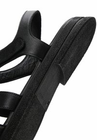 Sandale en cuir noir avec des sangles ajustables et une semelle épaisse texturée, présentée en gros plan sur un fond blanc.