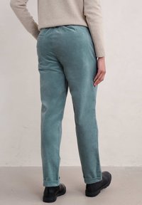 Osoba w turkusowych sztruksowych spodniach podwiniętych na nogawkach, beżowym swetrze i czarnych butach, stojąca na tle jasnej, gładkiej ściany.